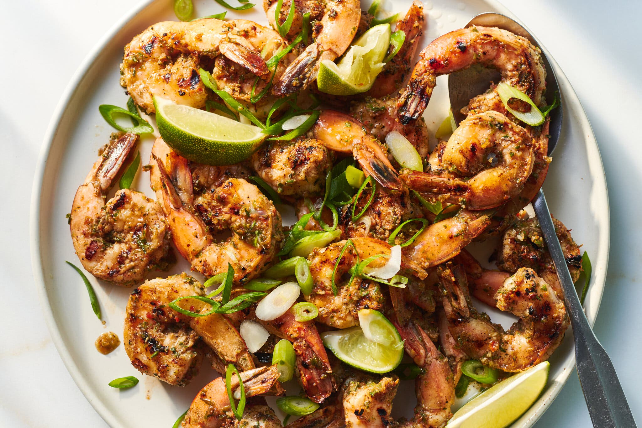A white plate is filled with shrimp flecked with spices, lime wedges and sliced scallions.
