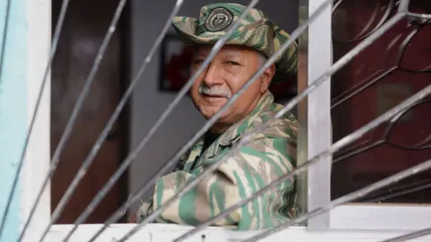 Nicole Kolster/BBC Edith Perales looks at the camera through a window. He is wearing a uniform in camouflage colours and a hat with the emblem of the militia. He has a grey moustache and greying temples.