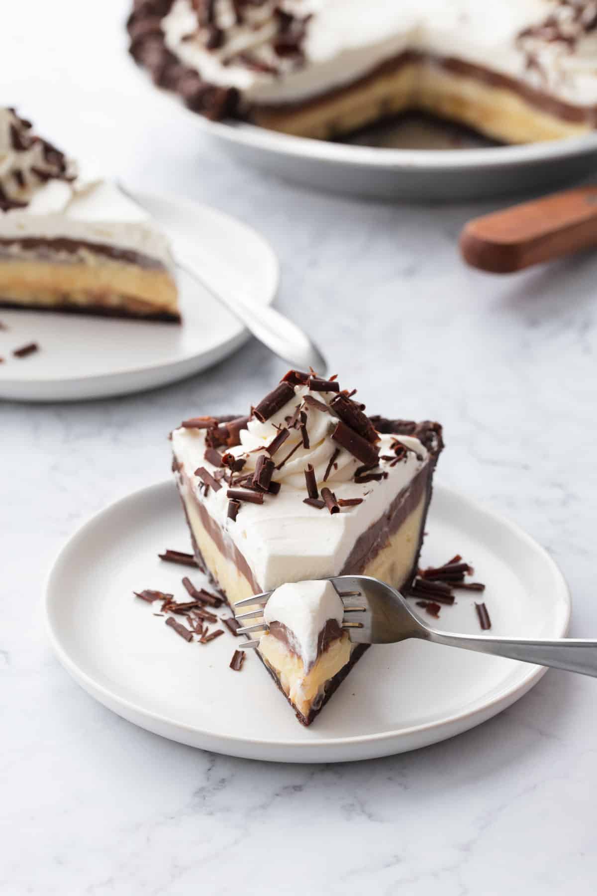 Fork cutting the tip off a slice of chocolate banana cream pie topped with whipped cream.