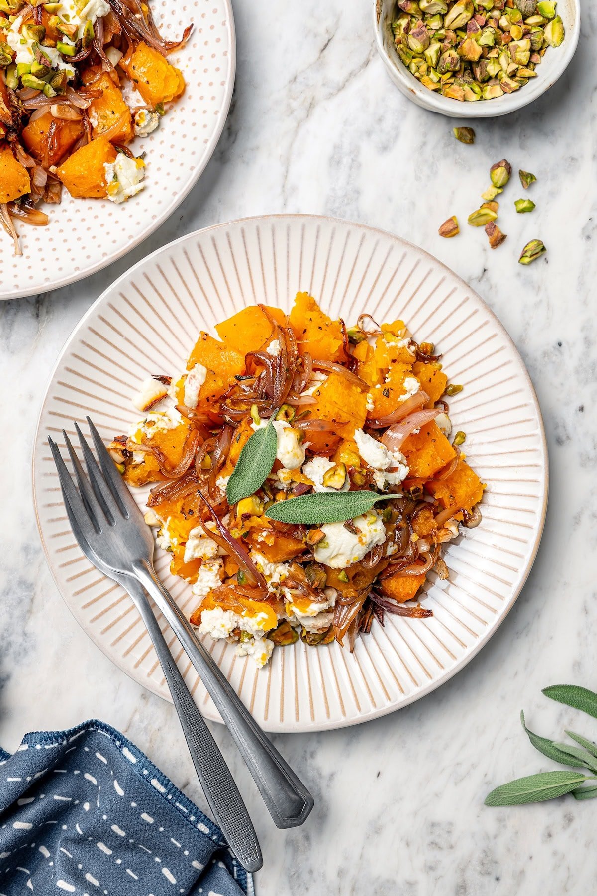 Baked butternut squash on 2 plates, topped with goat cheese, pistachios, shallots, and sage.