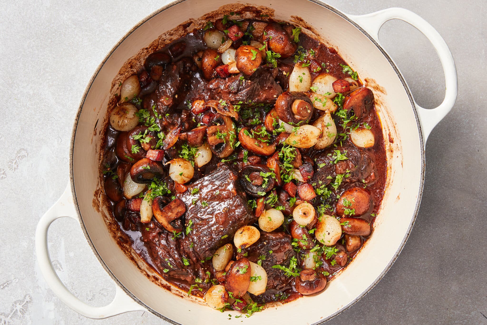 A white Dutch oven containing dark, wine-braised beef with whole pearl onions and sautéed mushrooms.
