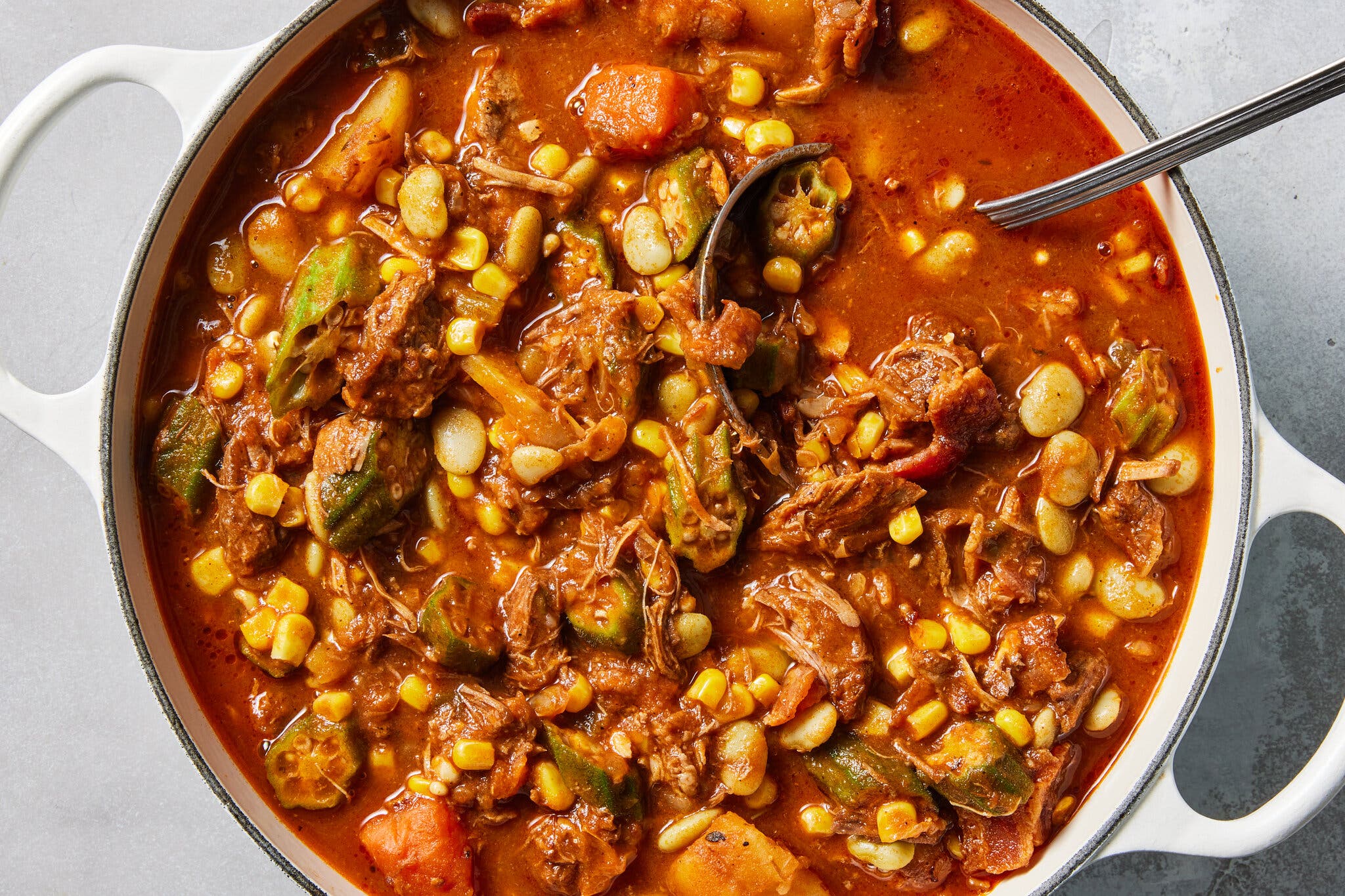 A silver ladle rests in a white pot of hearty stew featuring shredded beef, chicken, sliced green okra, yellow corn kernels and lima beans.