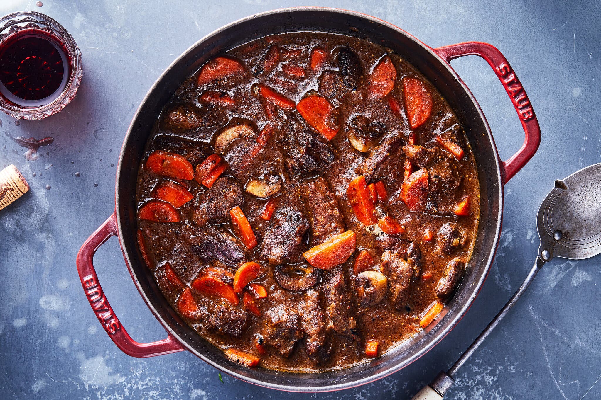 A deep red Staub Dutch oven filled with a glossy beef stew and bright orange half-moon carrot slices.