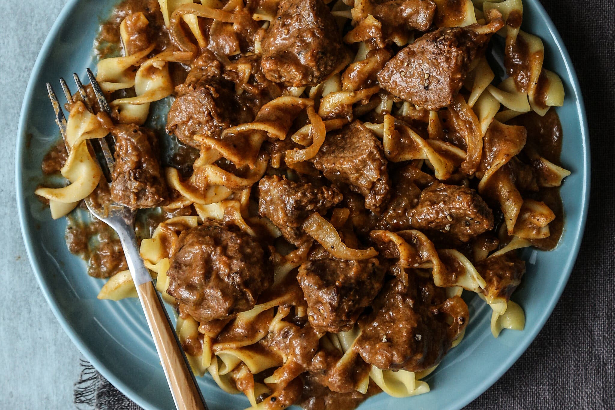 Tender chunks of beef coated in a rich brown gravy, served over a bed of buttery, wide spiral egg noodles on a light blue ceramic plate.